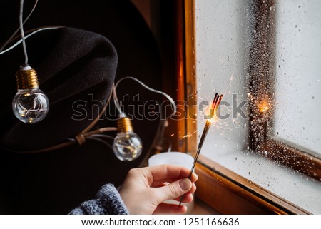 Winter concept. Hands girl holds Bengal lights on the background of a window, hat and decorated with led lights.