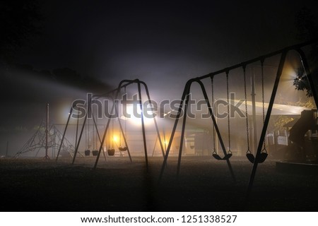 Swing sets backlit by streetlights on a dark foggy morning. Lake Benson Park in Garner North Carolina. Has a haunting or sci-fi feel.