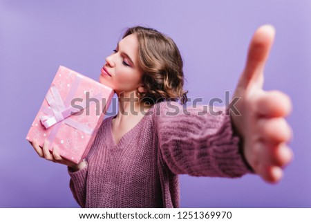 Indoor portrait of winsome girl with pink makeup holding present box. Photo of romantic curly lady with gift isolated on purple background.