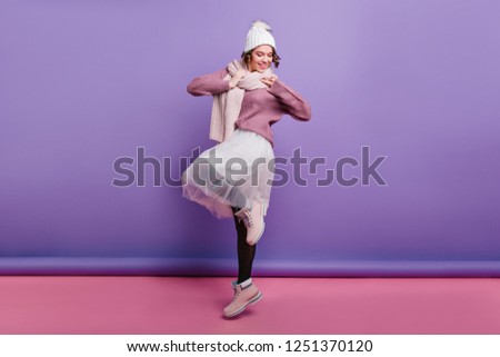 Full-length photo of winsome girl in long white skirt posing on one leg and laughing. Portrait of gorgeous female model in winter shoes and hat.