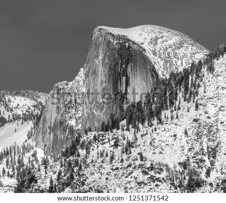Black and white picture of Half Dome, Yosemite national Park, covered in snow in the winter with trees on the shoulder of the slope and a dark sky above.