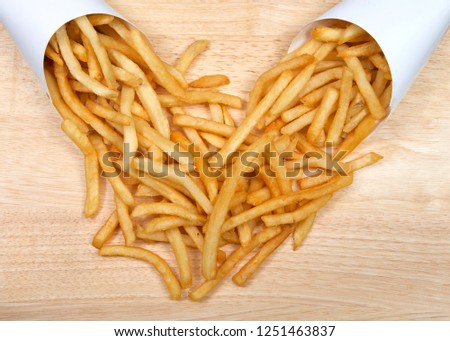 two white cups full of french fries spilling into each other on a light wood table. French fries have been popularized worldwide in large part by the large American fast food chains.