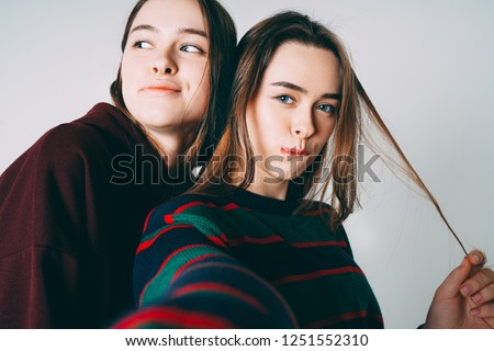 Two sisters twins beautiful girls in casual taking selfie on frontal camera of smartphone on grey background isolated