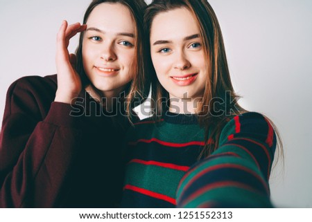 Two sisters twins beautiful girls in casual clothes taking selfie on frontal camera of smartphone on grey background isolated
