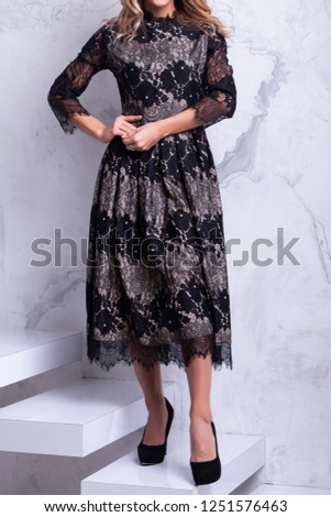 Young beautiful blond woman in a fashion black  dress posing on a white staircase, in the background a marble wall. Ideas of a New Year's dress for girls, New Year's fashion trends