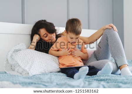 Little kid watching cartoons on tablet while sitting on the bed while his mother smiling and watching cartoons with him. Bedroom interior.