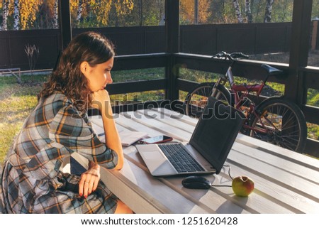 Young brunette woman sitting on outdoor terrace and reading unrecognizable text at laptop monitor. Mobile phone, eyeglasses and green apple are on wooden table. Online chatting, shopping concept.