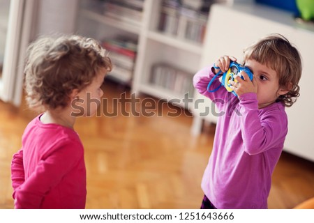 Little Girl Taking Photos Of Her Twin Sister With A Blue And Yellow Vintage Compact Camera In Living Room With Wooden Parquetry Flooring.