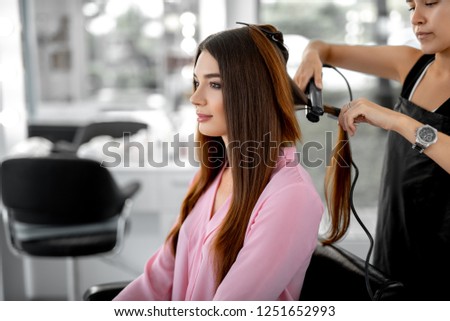 Waist up photo of attractive young woman looking in the mirror while female hairdresser styling her hair