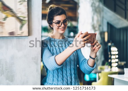 Positive young woman in stylish eyeglasses holding smartphone and smiling at front camera to make photo in social networks.Cheerful hipster girl taking selfie picture on cellular for blog in cafe