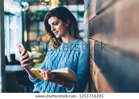 Attractive positive hipster student holding literature book while watching funny video on website on modern smartphone using 4G internet connection.Cheerful young woman reading good news on phone