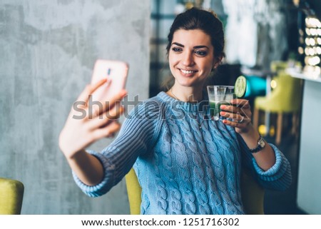 Cheerful young woman holding tasty cocktail in hand and making selfie photo on modern smartphone device.Positive hipster girl with beverage holding mobile phone and taking picture for internet blog