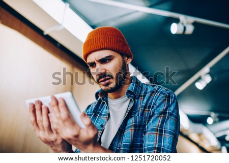 Angry hipster guy in stylish cap received email with bad news feeling unhappy from reading information via application on modern touch pad, millennial man holding digital tablet and watching video