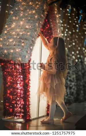 curly little girl sitting on present with white Christmas tree with smile and happy Christmas lights new year decoration near the window