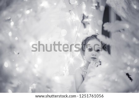 curly little girl sitting on present with white Christmas tree with smile and happy Christmas lights new year decoration