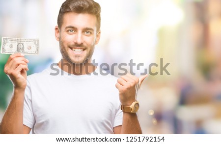 Young handsome man holding dollars over isolated background pointing and showing with thumb up to the side with happy face smiling