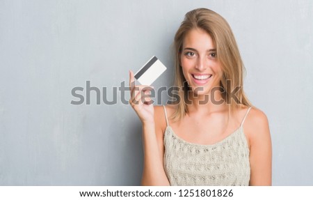 Beautiful young woman over grunge grey wall holding credit card with a happy face standing and smiling with a confident smile showing teeth