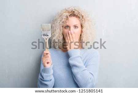 Young blonde woman with curly hair over grunge grey background holding paint brush cover mouth with hand shocked with shame for mistake, expression of fear, scared in silence, secret concept