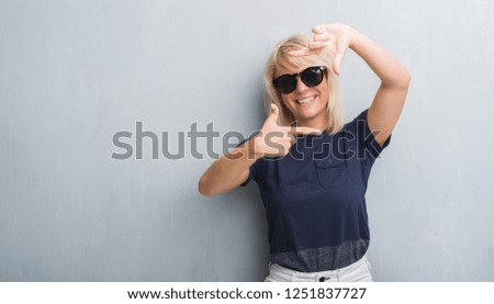 Adult caucasian woman over grunge grey wall wearing sunglasses smiling making frame with hands and fingers with happy face. Creativity and photography concept.