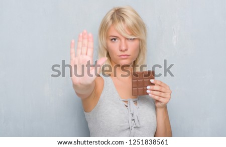 Caucasian adult woman over grey grunge wall eating chocolate bar with open hand doing stop sign with serious and confident expression, defense gesture