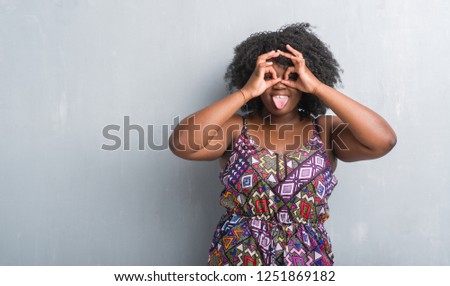 Young african american woman over grey grunge wall wearing colorful dress doing ok gesture like binoculars sticking tongue out, eyes looking through fingers. Crazy expression.