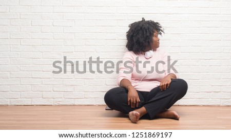 Young african american woman sitting on the floor wearing headphones looking to side, relax profile pose with natural face with confident smile.