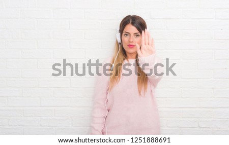 Young adult woman over white brick wall wearing headphones listening to music and dancing with open hand doing stop sign with serious and confident expression, defense gesture