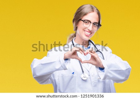 Beautiful young blonde doctor woman wearing medical uniform over isolated background smiling in love showing heart symbol and shape with hands. Romantic concept.