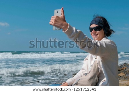 Woman makes selfie sitting by the sea on stone beach in autumn.