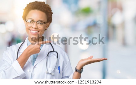 Young african american doctor woman wearing medical coat over isolated background amazed and smiling to the camera while presenting with hand and pointing with finger.