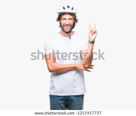 Handsome hispanic cyclist man wearing safety helmet over isolated background smiling with happy face winking at the camera doing victory sign. Number two.