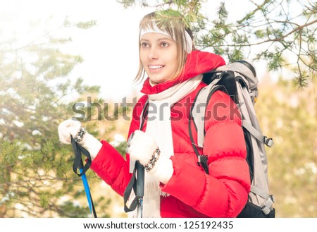 Beautiful girl in red jacket standing with backpack and ski poles. Happy smiling woman enjoys bright winter day. Winter snowy forest in background. Active sport and outdoor activity.