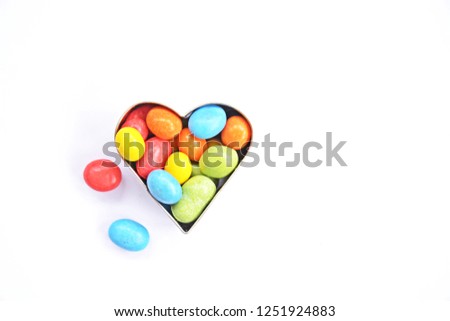 A cookie cutter in the shape of a heart lies on a white background and is filled with colorful chocolate lentils - Concept with sweet colorful chocolate lentils in Christmas shapes with space for text