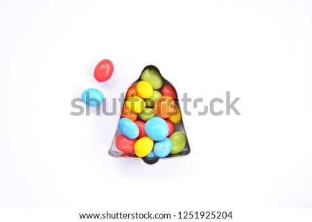 A cookie cutter in the shape of a bell lies on a white background and is filled with colorful chocolate lentils - Concept with sweet colorful chocolate lentils in Christmas shapes with space for text 