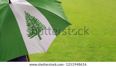 Norfolk Island flag umbrella. Close up of printed umbrella over green grass lawn background. Rainy weather forecast. Climate change and global warming concept.