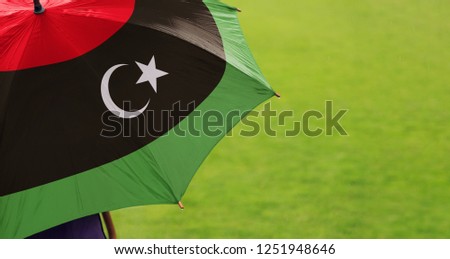 Libya flag umbrella. Close up of printed umbrella over green grass lawn background. Rainy weather forecast. Climate change and global warming concept.