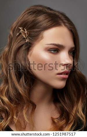 Close up portrait of a young brunette lady with natural make-up and wavy hairstyle. Her tresses are adorned with golden twig hair clip. The girl posing over the grey background, looking to the side. Royalty-Free Stock Photo #1251987517