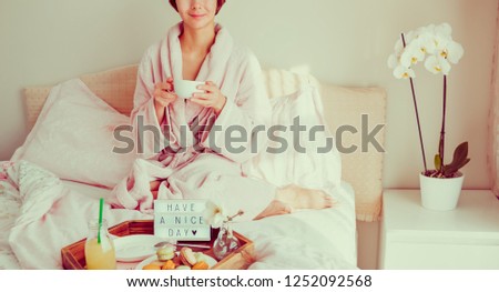 Good morning concept. Woman in bathrobe sitting on the bed, drinking coffee and has her breakfast in bed with Have a nice day text on lighted box. Hospitality, care, service. Wide banner. Copy space