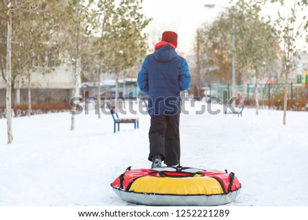 cute young boy in hat blue jacket plays with snow, has fun, smiles. Teenager goes with tube on hills in winter park. Active lifestyle, winter activity, outdoor winter games, snowballs. 