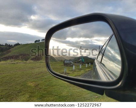 Side view mirror of a car. Reflection of woman viewing sun rays and sunset. Travel road trip image. Beautiful landscape image from car mirror. Weekend break road trip. Sightseeing car journey.