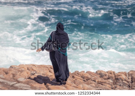Arabic woman in traditional clothes looking at big waves in Hermanus, South Africa