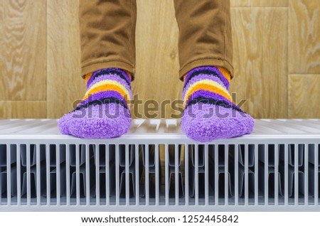 View from above of a woman's feet in bright warm winter socks warming them near a heating radiator at home. Part of body, selective focus.