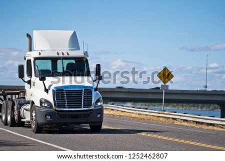 White powerful big rig day cab with roof spoiler local hauler semi truck with flat bed semi trailer driving on the road along the river with bridge across the river to deliver commercial cargo Royalty-Free Stock Photo #1252462807