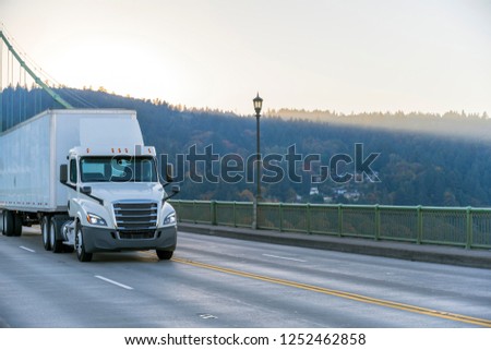 Modern white day cab big rig semi truck with roof spoiler for reduce air resistance transporting commercial cargo in dry van semi trailer driving on the St Johns bridge Royalty-Free Stock Photo #1252462858