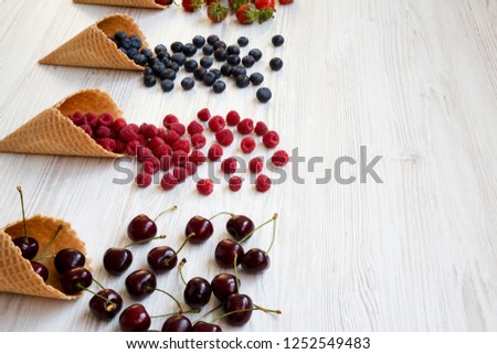 Waffle sweet ice cream cones with raspberries, cherries, strawberries and blueberries over white wooden background, side view. Copy space.