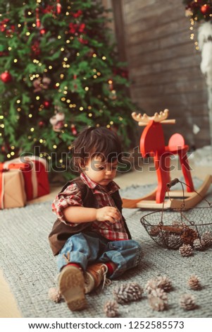 Small boy is sitting on carpet and playing with conelet in Christmas interior, red elements, pine cones, Christmas tree