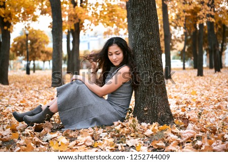 Pretty woman is sitting in autumn park near big tree. Beautiful landscape at fall season.