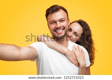 Young smiling man and woman in embrace on orange background looking at camera taking selfie