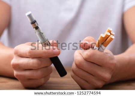 Man Holding Vape And Tobacco Cigarette Over Desk Royalty-Free Stock Photo #1252774594