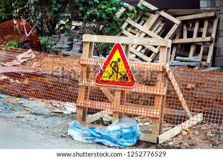 The fence of the grid and a wooden pallet with a warning sign about the repair work in front of the pit.